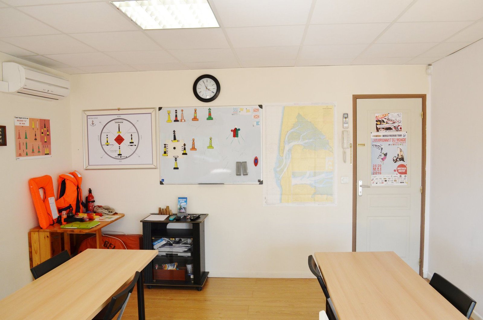 Une salle de classe avec des tables, des chaises, un tableau blanc et une horloge au mur.